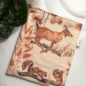 Può includere: Una custodia in tessuto beige con un motivo boschivo che mostra un cervo, scoiattoli e fogliame autunnale nei toni del marrone e dell'arancione. La custodia è rettangolare con un interno arancione.