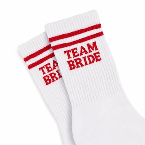 Könnte beinhalten: Ein Paar weiße Crew-Socken mit zwei horizontalen roten Streifen am oberen Bündchen. Der Schriftzug "TEAM BRIDE" ist in roten Blockbuchstaben auf der Außenseite jeder Socke direkt unter den roten Streifen auf dem gerippten Stoff aufgedruckt.