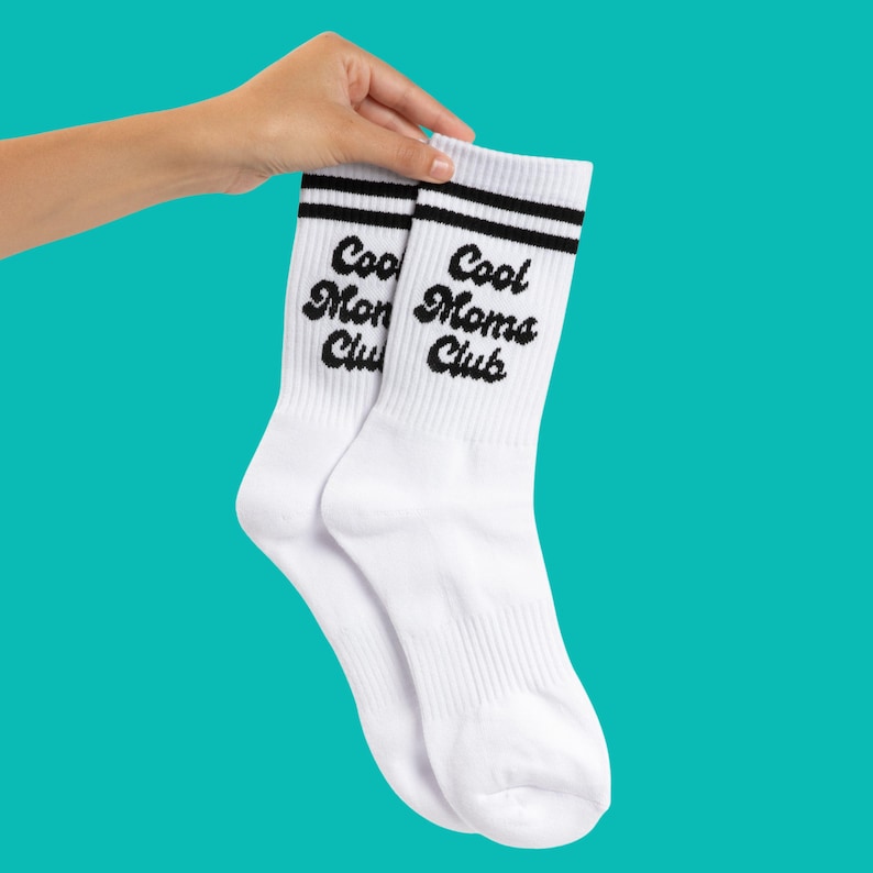May include: A pair of white socks with black stripes at the top. The socks have the words "Cool Moms Club" printed in a black script font. The socks are held up by a hand against a teal background.