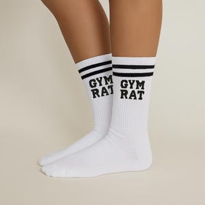 K&ouml;nnte beinhalten: Wei&szlig;e Sportsocken mit schwarzen Streifen am oberen Rand und dem schwarzen Aufdruck "GYM RAT" an der Seite. Die Socken sind gerippt und werden an den Beinen einer Person getragen.