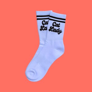 Könnte beinhalten: Ein Paar weiße Socken mit schwarzen Streifen und dem Schriftzug "Cat La Lady" in schwarzer Schrift. Die Socken sind vor einem einfarbigen orangefarbenen Hintergrund abgebildet.
