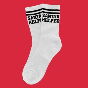 Könnte beinhalten: Paar weiße Sportsocken mit schwarzen Streifen am oberen Rand. Die Worte "SANTA'S HELPER" sind in Schwarz auf den Sockenbündchen aufgedruckt. Die Socken werden vor einem roten Hintergrund gezeigt.