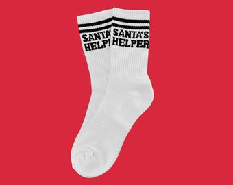 Puede incluir: Par de calcetines deportivos blancos con rayas negras en la parte superior. Las palabras "SANTA'S HELPER" están impresas en negro en los puños de los calcetines. Los calcetines se muestran sobre un fondo rojo liso.