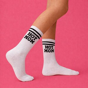 May include: White athletic socks with black stripes and the words "HOT MOM" in black block letters. The socks are ribbed and worn on legs against a pink background.