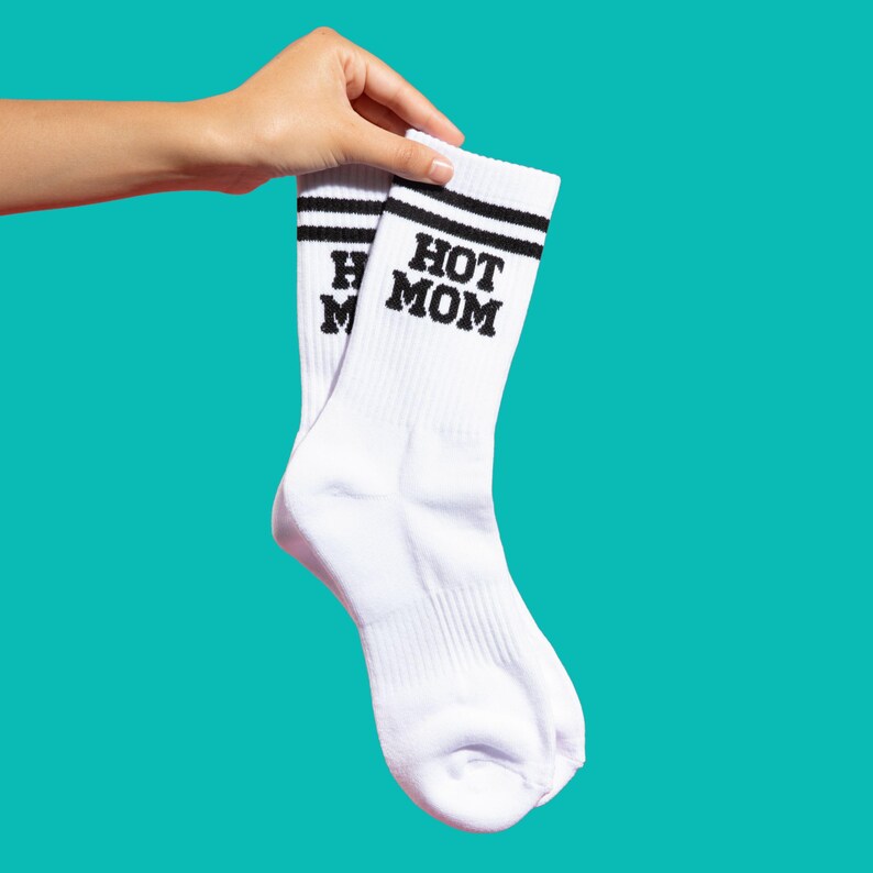 May include: A pair of white socks with black stripes at the top and the words "HOT MOM" printed in black. The socks are held up against a teal background.