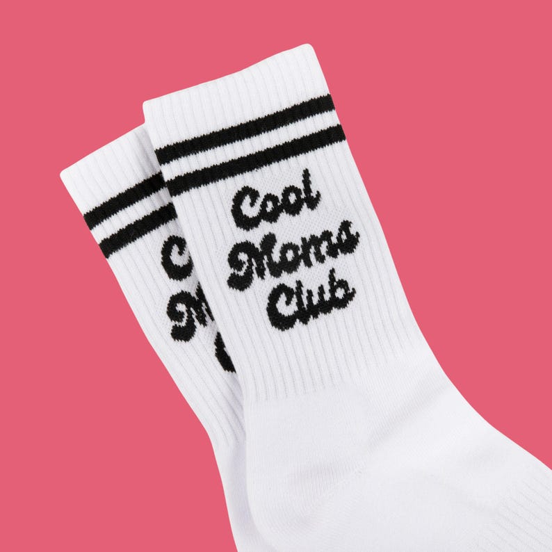 May include: White socks with black stripes and the text "Cool Moms Club" in a script font. The socks are on a pink background. These socks are a fun and stylish accessory.