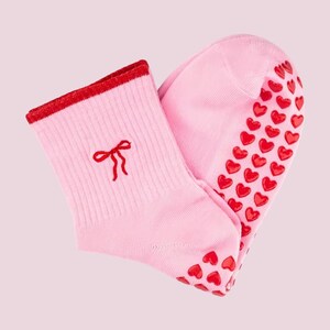 Puede incluir: Un par de calcetines rosas con un lazo rojo bordado cerca del pu&ntilde;o. El pu&ntilde;o est&aacute; ribeteado en rojo y las suelas est&aacute;n cubiertas de agarres rojos en forma de coraz&oacute;n. Los calcetines est&aacute;n hechos de un material suave y acanalado.