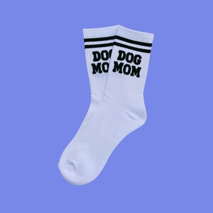 Könnte beinhalten: Ein Paar weiße Socken mit schwarzen Streifen am oberen Rand. Die Socken haben die Worte "DO DOG MO MOM" in schwarzen Blockbuchstaben aufgedruckt. Der Hintergrund ist helllila.