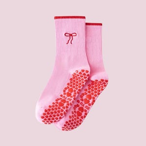 Puede incluir: Un par de calcetines rosas con detalles rojos. Los calcetines tienen un lazo rojo bordado cerca de la parte superior y un patr&oacute;n de corazones rojos en las suelas. Los calcetines est&aacute;n hechos de un material suave.