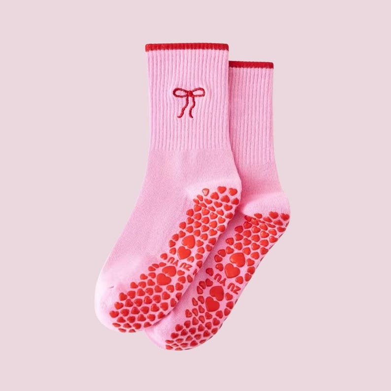 Puede incluir: Un par de calcetines rosas con detalles rojos. Los calcetines tienen un lazo rojo bordado cerca de la parte superior y un patr&oacute;n de corazones rojos en las suelas. Los calcetines est&aacute;n hechos de un material suave.