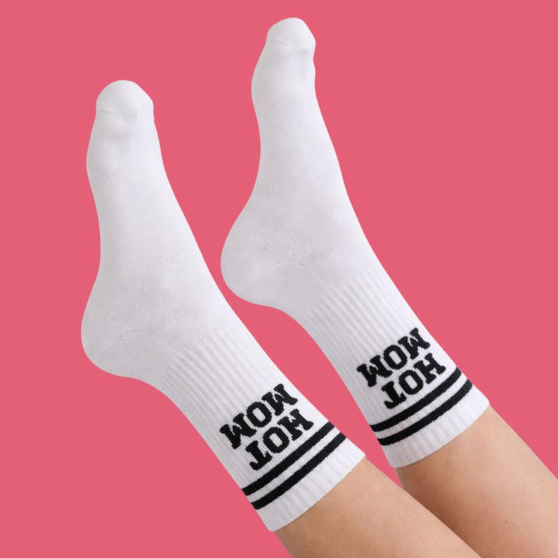 May include: White socks with the text "HOT MOM" in black, with two black stripes at the top. The socks are ribbed and are against a pink background.