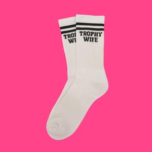 Könnte beinhalten: Ein Paar weiße Socken mit schwarzen Streifen oben. Der Text "TROPHY WIFE" ist in Schwarz auf die Socken gedruckt. Die Socken werden vor einem hellrosa Hintergrund präsentiert.