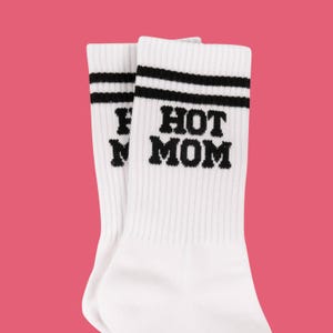 May include: A pair of white socks with black stripes at the top and the words "HOT MOM" in bold black letters. The socks are ribbed and have a pink background.