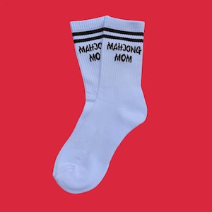 Könnte beinhalten: Ein Paar weiße Socken mit schwarzen Streifen am oberen Rand. Der Text "MAHJONG MOM" ist in Schwarz auf die Socken gedruckt. Die Socken werden vor einem roten Hintergrund präsentiert.