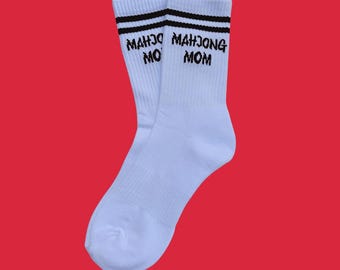 Puede incluir: Un par de calcetines blancos con rayas negras en la parte superior. El texto "MAHJONG MOM" está impreso en negro en los calcetines. Los calcetines se muestran sobre un fondo rojo.