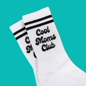 May include: A pair of white socks with black stripes at the top. The words "Cool Moms Club" are printed in black script on the side of the socks. The socks are on a teal background.