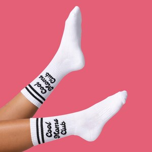 May include: White athletic socks with black stripes and the text "Cool Moms Club" in a script font. The socks are displayed against a pink background.
