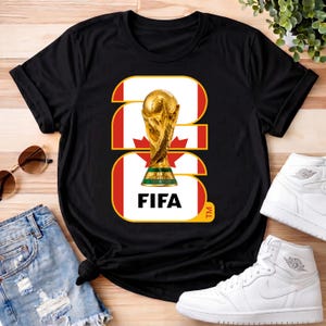 Może przedstawiać: Czarna koszulka z złotą grafiką trofeum FIFA World Cup. Projekt zawiera kanadyjską flagę i słowo "FIFA". Koszulka jest prezentowana z okularami przeciwsłonecznymi, dżinsowymi szortami i białymi trampkami.