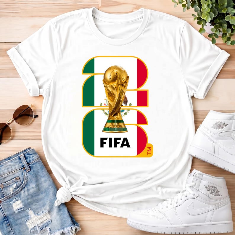 Może przedstawiać: Biała koszulka z złotym trofeum i napisem "FIFA". Projekt zawiera kolory meksykańskiej flagi. Casualowy element garderoby, odpowiedni dla fan&oacute;w sportu.