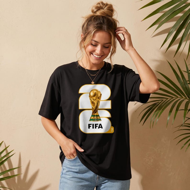 Może przedstawiać: Czarny t-shirt z złotą grafiką trofeum FIFA World Cup. Projekt zawiera słowo "FIFA" w białym prostokącie ze złotą obw&oacute;dką. Koszulka jest noszona przez osobę z blond włosami.