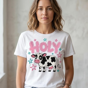May include: White t-shirt with a cartoon cow graphic. The cow is black and white with a pink udder. The word "HOLY" is in pink bubble letters above the cow, surrounded by pink and teal stars.