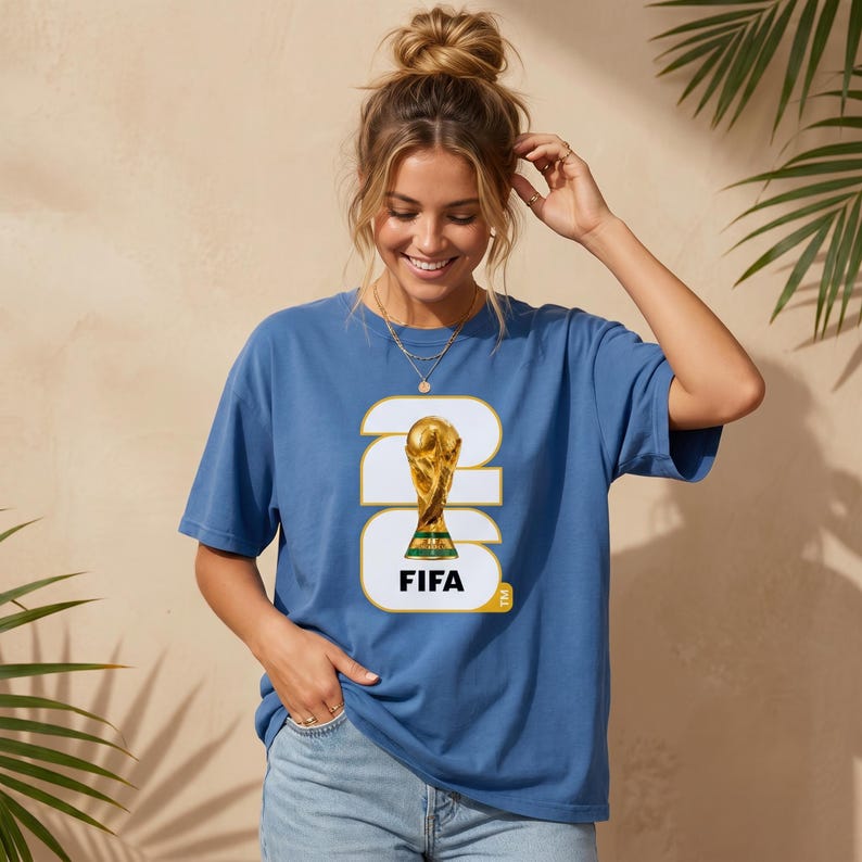 Może przedstawiać: Niebieska koszulka z grafiką trofeum Mistrzostw Świata FIFA. Projekt zawiera liczbę 26 i słowo "FIFA". Koszulka jest w jednolitym niebieskim kolorze.