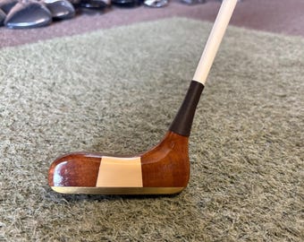 May include: A vintage wooden golf club with a polished, brown head and a white shaft. The club head features a light-colored stripe and a brass-colored sole. The grip is wrapped in a dark, textured material. The club is designed for golf.