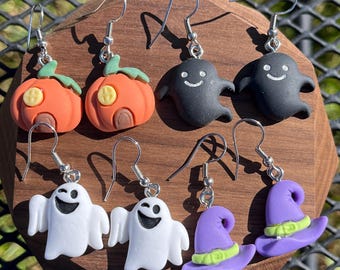May include: Four pairs of dangle earrings featuring miniature charms: orange pumpkins with green stems, black ghosts with white smiles, white ghosts with black open mouths, and purple witch hats with light green bands, all on silver-toned hooks.