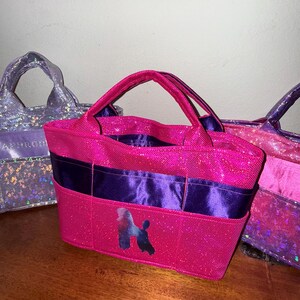 以下が含まれることがあります： ピンクと紫の色合いの、キラキラしたトートバッグが3つ。中央のバッグは鮮やかなピンク色で、紫のリボンとプードルのシルエットが特徴です。他の2つのバッグは、ピンクと紫を混ぜたもので、キラキラした仕上がりになっています。バッグには持ち手が付いています。
