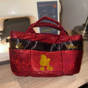 以下が含まれることがあります： テクスチャード加工のパターン、黒いサテンのトリム、そして「Falee」と「Poodles」と書かれた金色の文字が特徴の赤いハンドバッグ。前面には金色のプードルのシルエットがあります。バッグにはハンドルと複数のポケットが付いています。