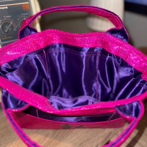 以下が含まれることがあります： 鮮やかなピンクのスパンコールが施された外側と、濃い紫色のサテンの内側が特徴の手作りのトートバッグ。バッグにはお揃いのピンクのハンドルと構造化されたデザインが施されています。内側には複数のポケットがあります。