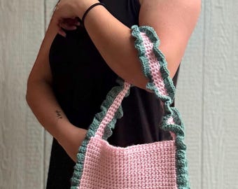 May include: A handmade pink crochet bag with a green ruffled trim. The bag has a rectangular shape and a matching strap with the same green ruffle detail. The bag is held by a person wearing a black dress.