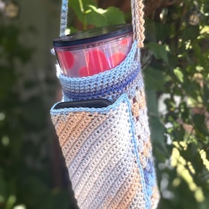 May include: Crocheted water bottle holder in shades of blue, tan, and white, with a matching strap. The holder has a pocket for a phone or other small items. A clear tumbler with a black lid and red polka dots is inside the holder.