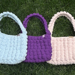 May include: Three handmade crochet bags in pastel colors: light blue, purple, and pink. Each bag features a textured, bubble-like stitch pattern and matching handles. The bags are displayed on a bed of green grass.