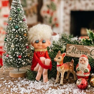 May include: A vintage-style Christmas display featuring a small flocked bottle brush tree with colorful ornaments, a doll with white hair in a red outfit, a small deer figurine, a Santa Claus figure, and a sign reading Merry Christmas.
