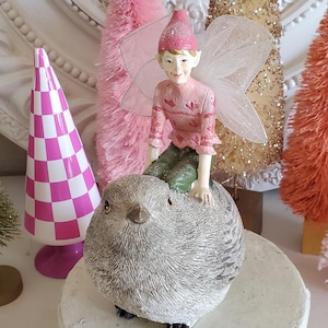 Puede incluir: Una figura caprichosa de un hada montada en un pájaro gris, posada sobre un pedestal blanco. El hada viste un atuendo rosa y tiene delicadas alas. En el fondo, árboles decorativos rosas y dorados con forma de cepillo.
