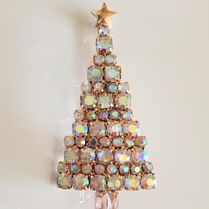 May include: A Christmas tree brooch with a gold-tone metal frame and iridescent, square-cut rhinestones. The tree is topped with a gold star and has a rectangular base. A festive holiday accessory.