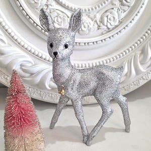 May include: A silver glitter-covered deer figurine with a small gold bell hanging from a cord around its neck, positioned next to a miniature bottle brush tree that transitions from a pink top to a cream-colored base.