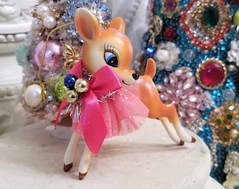 May include: A small deer figurine with a tan body, white underbelly, and large blue eyes. The deer wears a bright pink satin bow and a pink tulle skirt with silver glitter accents. Small blue and gold spherical ornaments are attached to the bow.
