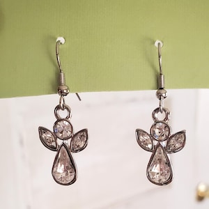 Puede incluir: Un par de pendientes de &aacute;ngel plateados con detalles de cristal transparente. Cada pendiente presenta un cuerpo de cristal en forma de l&aacute;grima, dos cristales en forma de ala y una cabeza de cristal circular. Los pendientes cuelgan de un gancho plateado.