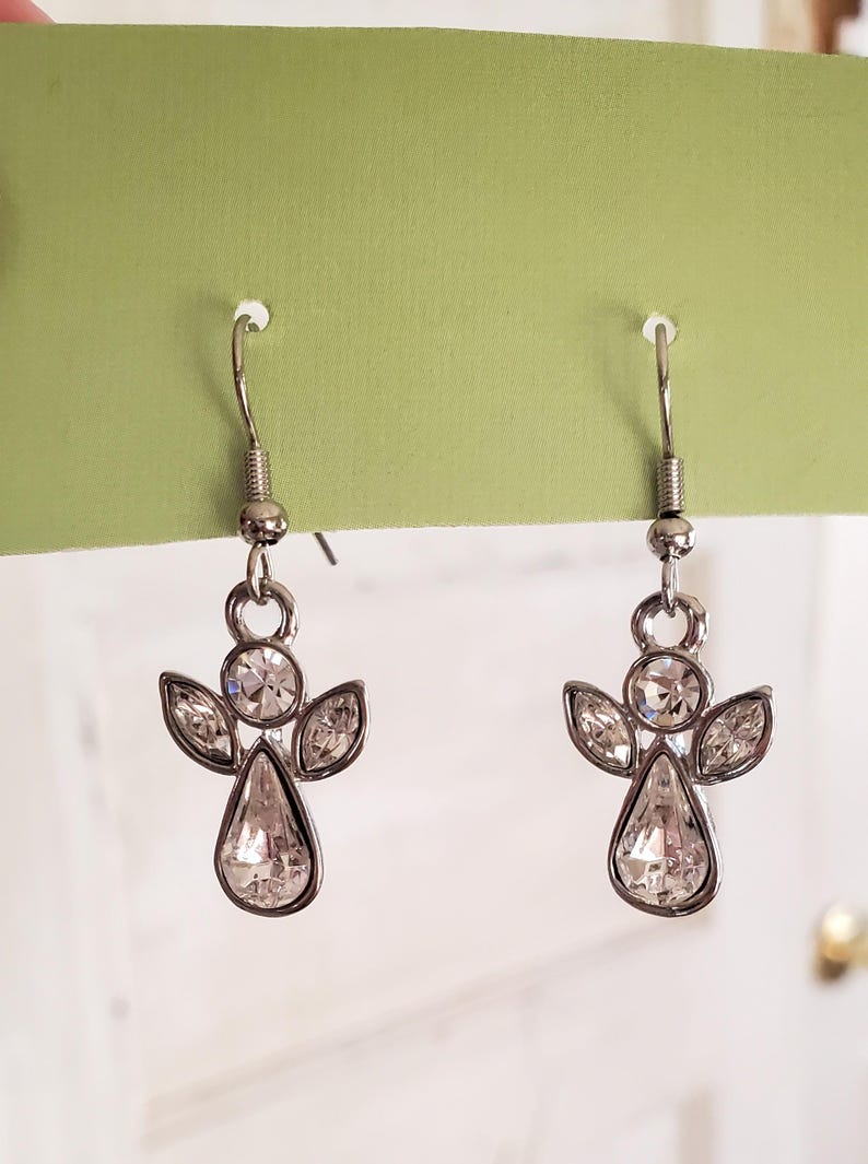 Puede incluir: Un par de pendientes de &aacute;ngel plateados con detalles de cristal transparente. Cada pendiente presenta un cuerpo de cristal en forma de l&aacute;grima, dos cristales en forma de ala y una cabeza de cristal circular. Los pendientes cuelgan de un gancho plateado.