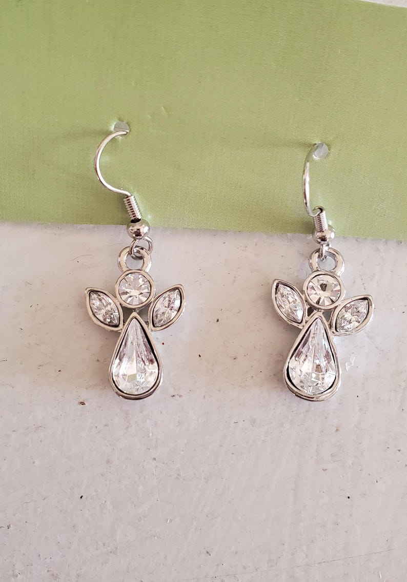 Puede incluir: Un par de pendientes plateados con forma de &aacute;ngel. Cada pendiente presenta un cuerpo de cristal transparente en forma de l&aacute;grima, con tres cristales m&aacute;s peque&ntilde;os que forman las alas. Un cristal redondo se encuentra en la cabeza y un gancho plateado completa el dise&ntilde;o.
