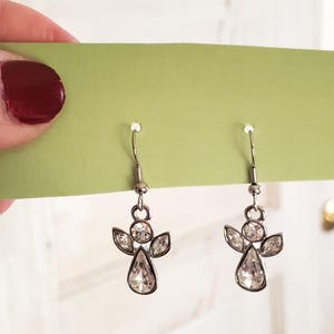 Puede incluir: Un par de pendientes de &aacute;ngel plateados con detalles de cristal transparente. Cada pendiente presenta un cuerpo de cristal en forma de l&aacute;grima, con cristales m&aacute;s peque&ntilde;os que forman alas y un halo. Los pendientes se exhiben en una tarjeta verde claro.
