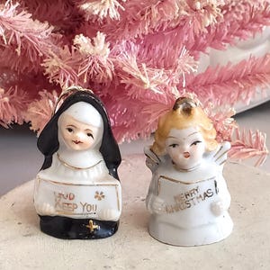 Puede incluir: Dos peque&ntilde;as figuras de cer&aacute;mica: una monja y un &aacute;ngel. La monja lleva un h&aacute;bito negro y sostiene un letrero que dice "God Keep You". El &aacute;ngel, con cabello rubio y alas, sostiene un letrero que dice "Merry Christmas". Ambos tienen detalles dorados.