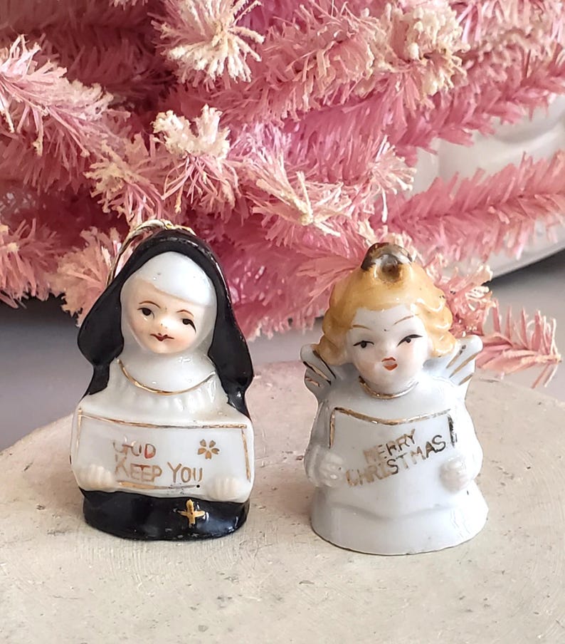 Puede incluir: Dos peque&ntilde;as figuras de cer&aacute;mica: una monja y un &aacute;ngel. La monja lleva un h&aacute;bito negro y sostiene un letrero que dice "God Keep You". El &aacute;ngel, con cabello rubio y alas, sostiene un letrero que dice "Merry Christmas". Ambos tienen detalles dorados.