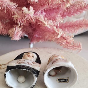 Puede incluir: Dos adornos de campana de cer&aacute;mica con caras pintadas y texto. Una campana dice "GOD KEEP YOU" y la otra "MERRY CHRISTMAS". Las campanas son blancas con detalles en negro y dorado. Un &aacute;rbol de Navidad rosa artificial est&aacute; en el fondo.