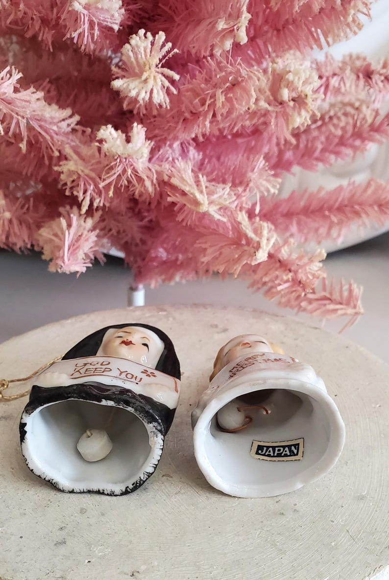 Puede incluir: Dos adornos de campana de cer&aacute;mica con caras pintadas y texto. Una campana dice "GOD KEEP YOU" y la otra "MERRY CHRISTMAS". Las campanas son blancas con detalles en negro y dorado. Un &aacute;rbol de Navidad rosa artificial est&aacute; en el fondo.