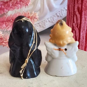 Puede incluir: Dos peque&ntilde;as figuras de cer&aacute;mica: una figura vestida de negro con una cuerda y un &aacute;ngel blanco con pelo rubio y alas. El &aacute;ngel tiene un detalle de cuerda naranja. Ambas figuras est&aacute;n sobre una superficie de color claro.