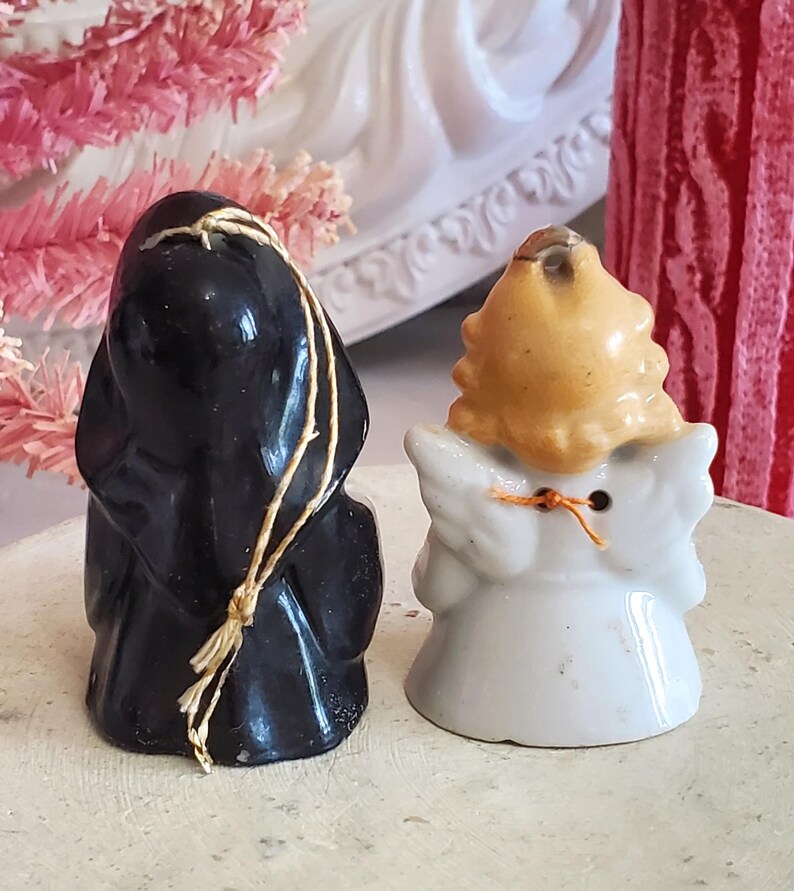 Puede incluir: Dos peque&ntilde;as figuras de cer&aacute;mica: una figura vestida de negro con una cuerda y un &aacute;ngel blanco con pelo rubio y alas. El &aacute;ngel tiene un detalle de cuerda naranja. Ambas figuras est&aacute;n sobre una superficie de color claro.