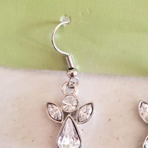 Puede incluir: Pendientes de &aacute;ngel plateados con cristales transparentes y brillantes. Cada pendiente presenta un cristal en forma de l&aacute;grima en el centro, rodeado de cristales m&aacute;s peque&ntilde;os con forma de marquesa y un cristal redondo en la parte superior. Cierre de gancho.